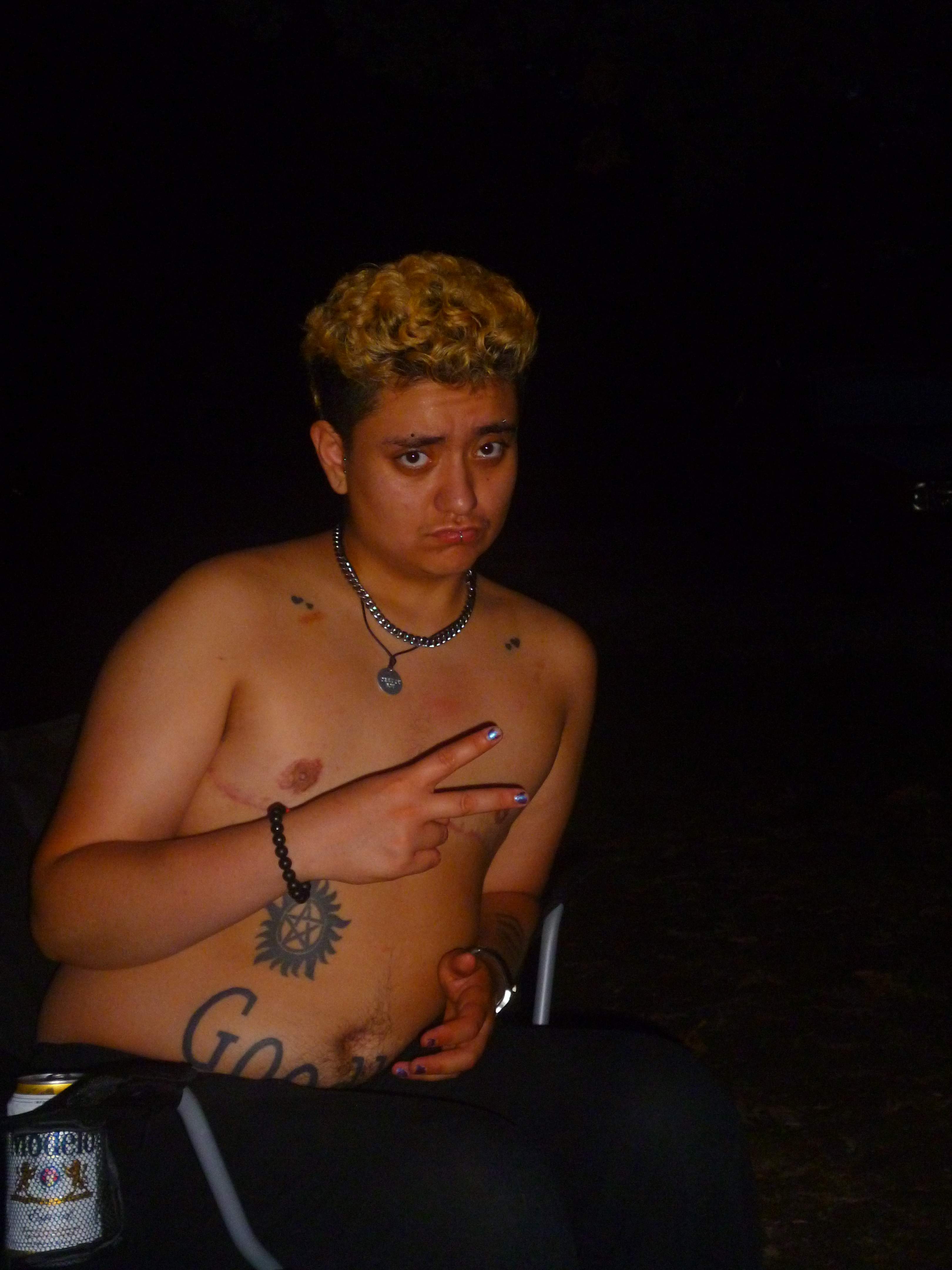 David, shirtless, in the dark in a folding camp chair holding up a peace sign.
